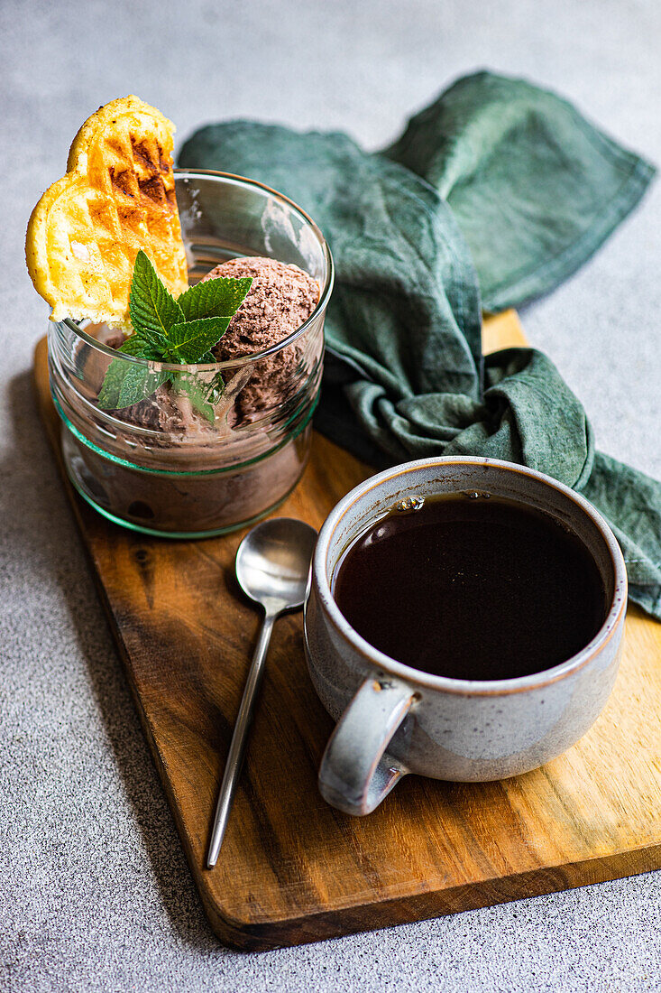 High angle of glass of homemade coffee ice cream with waffle and fresh mint near spoon and cup of coffee placed on wooden tray with napkin against gray surface