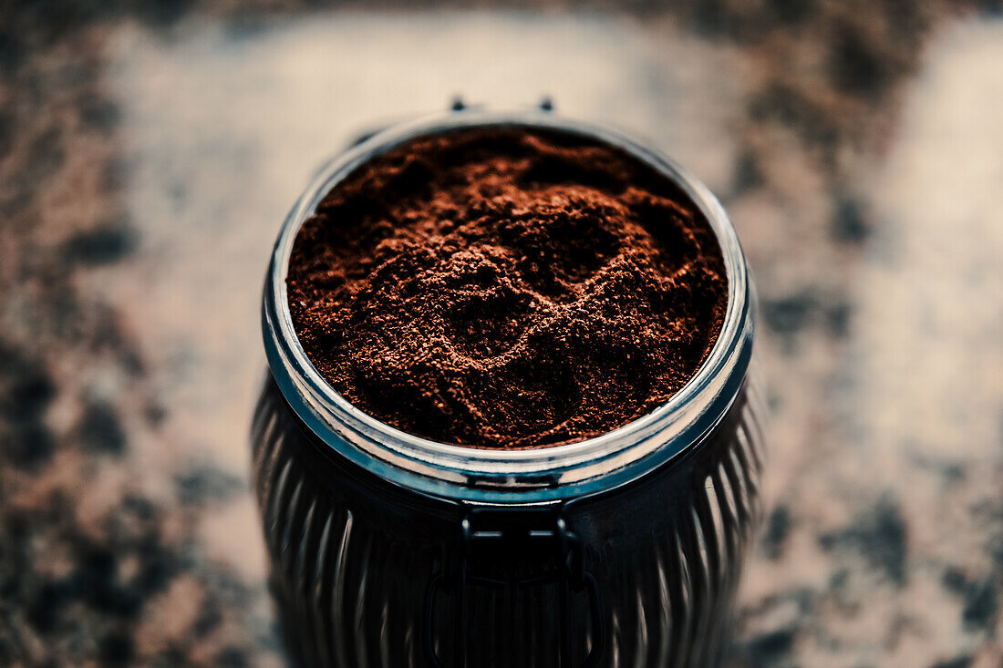Draufsicht auf fein gemahlenen Kaffee in einem gerippten Glasgefäß, das seine reiche Textur und Tiefe hervorhebt, vor einem gesprenkelten Hintergrund