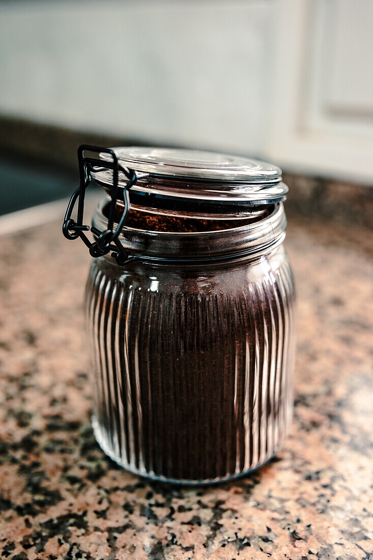 Nahaufnahme eines gerippten Glasgefäßes mit frisch gemahlenem Kaffee, das mit einem Klemmdeckel aus Metall auf einer gesprenkelten Granitarbeitsplatte steht