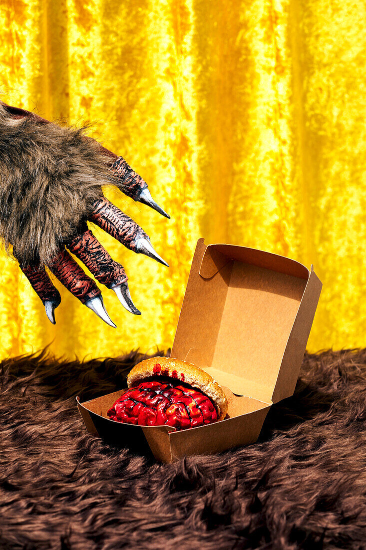 Hairy paw with metal claws of anonymous monster grabbing bloody brain with burger bun from paper box placed on fur blanket against golden curtain