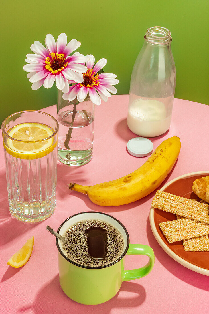 Banane neben Glasflasche mit Milch und Wasser mit Zitronenscheibe vor Blumen in Vase und Tasse mit aromatischem Kaffee auf rosa Tisch vor grünem Hintergrund