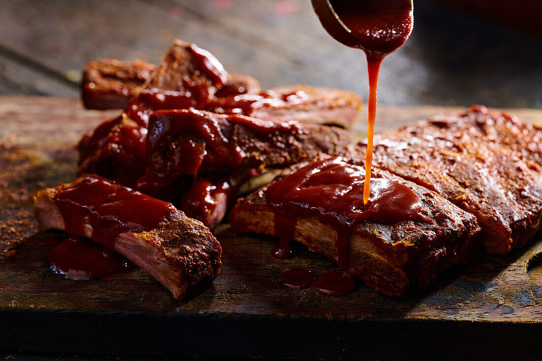 High angle of anonymous cook pouring barbecue on grilled ribs placed on wooden chopping board against blurred background
