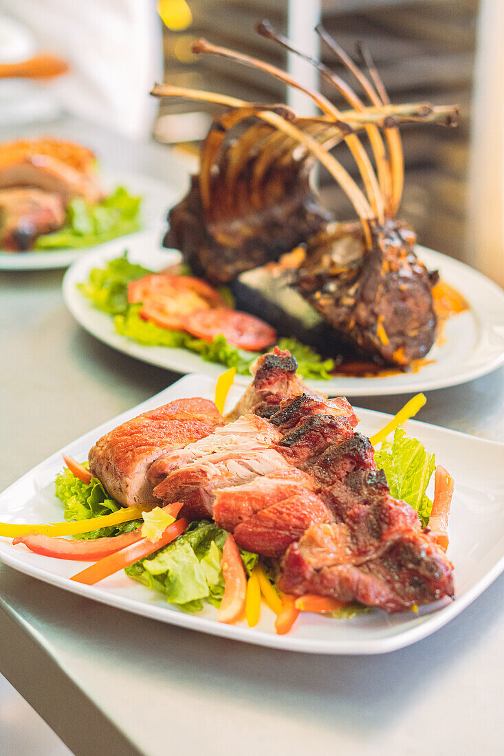 High angle of appetizing pieces of meat served on plate with pepper and lettuce leaves near rack of lamb with tomato slices in restaurant kitchen