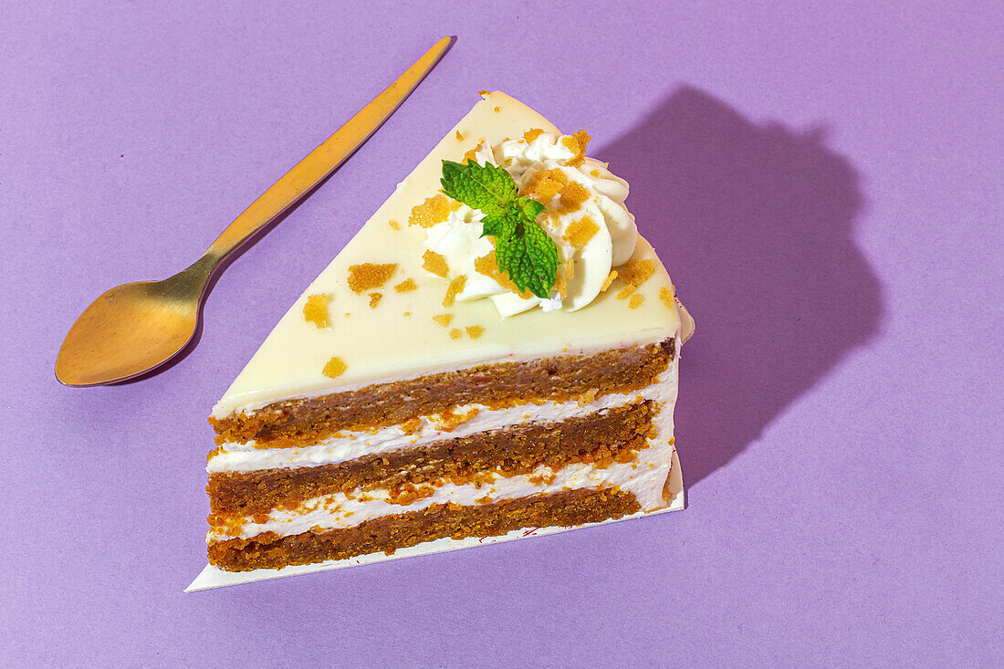 From above close up of slice of tasty sweet carrot sponge cake with cream decorated with mint leaf served on plate with spoon on table on purple background