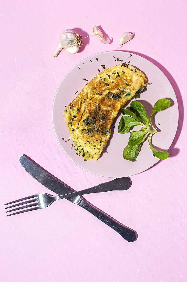 Tasty omelette on plate against fresh parsley sprigs with garlic cloves on pink background