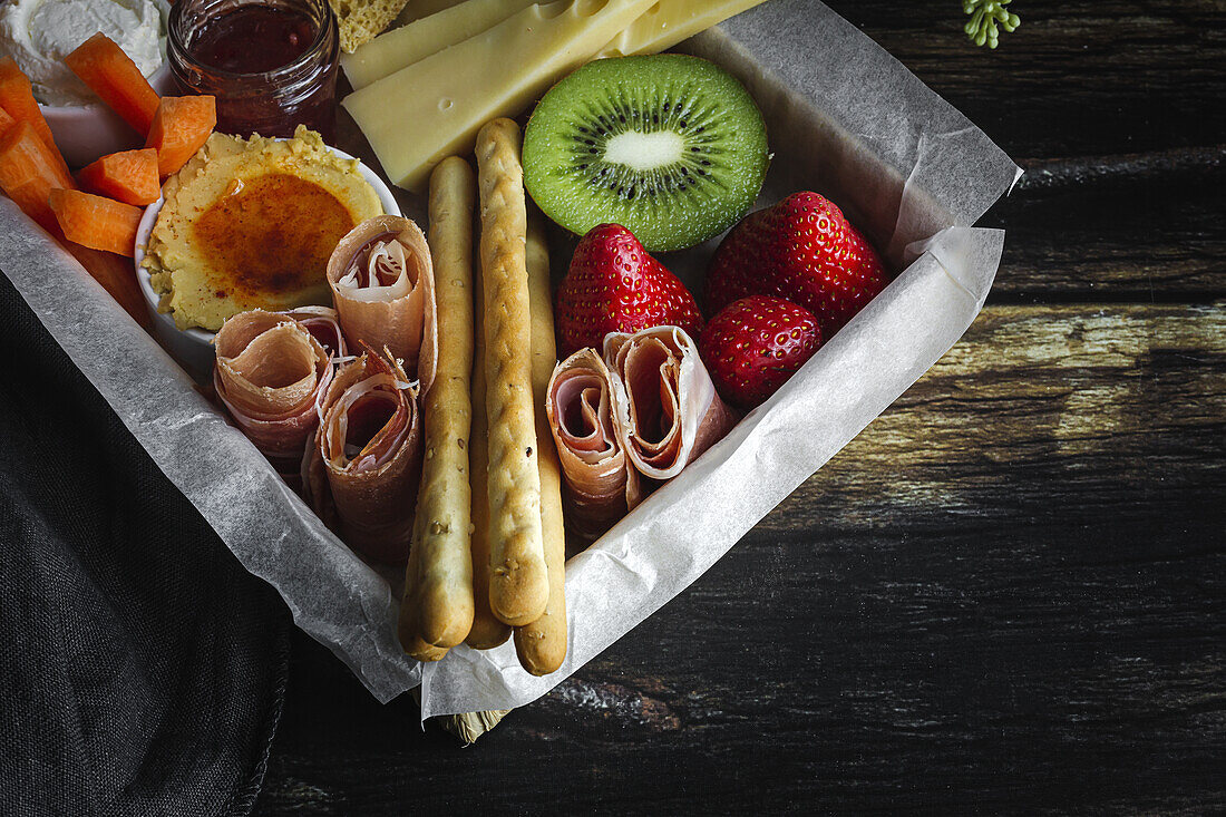 Von oben Brunch-Box mit verschiedenen Wurst- und Käsesorten und Knäckebrot in der Nähe von reifen Kiwis, süßen Erdbeeren und geschälten Mandarinen in der Nähe von Marmelade in einem Glas auf einem Holztisch in der Nähe einer Serviette