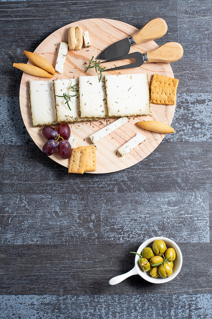 Top view of appetizing cheese served on wooden table with ripe grapes and crackers decorated by rosemary sprigs near olives in bowls on table