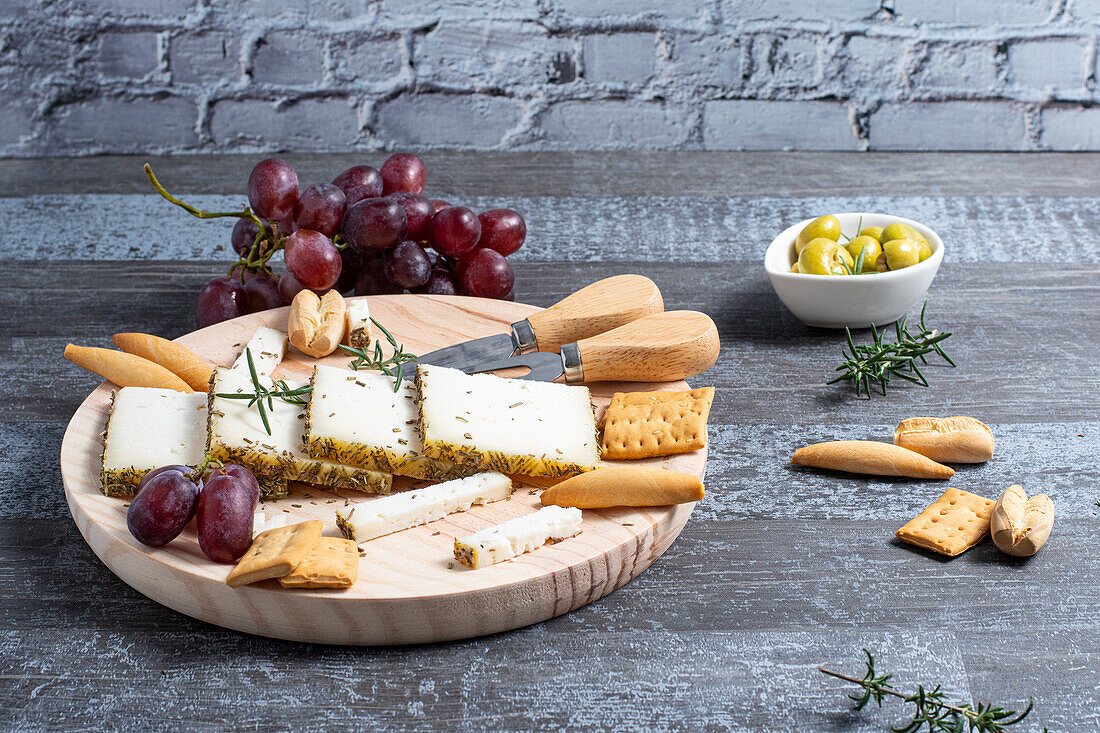 Appetizing cheese served on wooden table with ripe grapes and crackers decorated by rosemary sprigs near olives in bowls on table