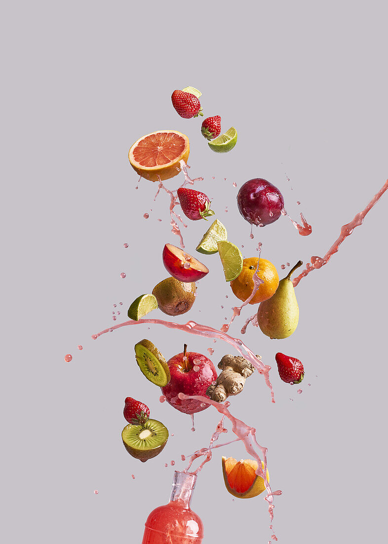 Strauß verschiedener frischer Früchte und Ingwer fällt in Glasflasche mit gesundem Saft vor grauem Hintergrund