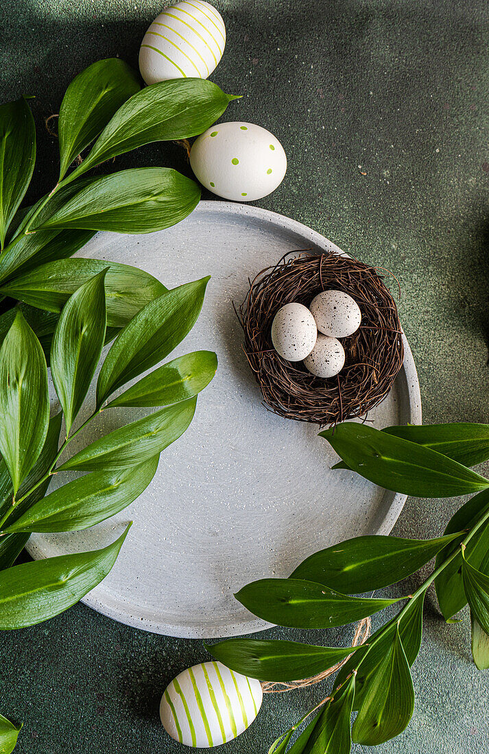 Von oben gedeckter Ostertisch mit Keramiktellern und Ostereiern in einem Nest, umgeben von italienischem Ruscus und Polsterblättern des Schleierkrauts