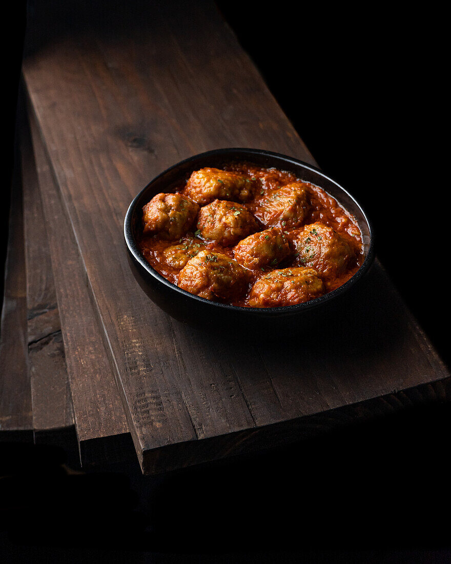From above of black bowl with delicious meatballs in tomato sauce with herbs placed on pile of wooden boards in dark room