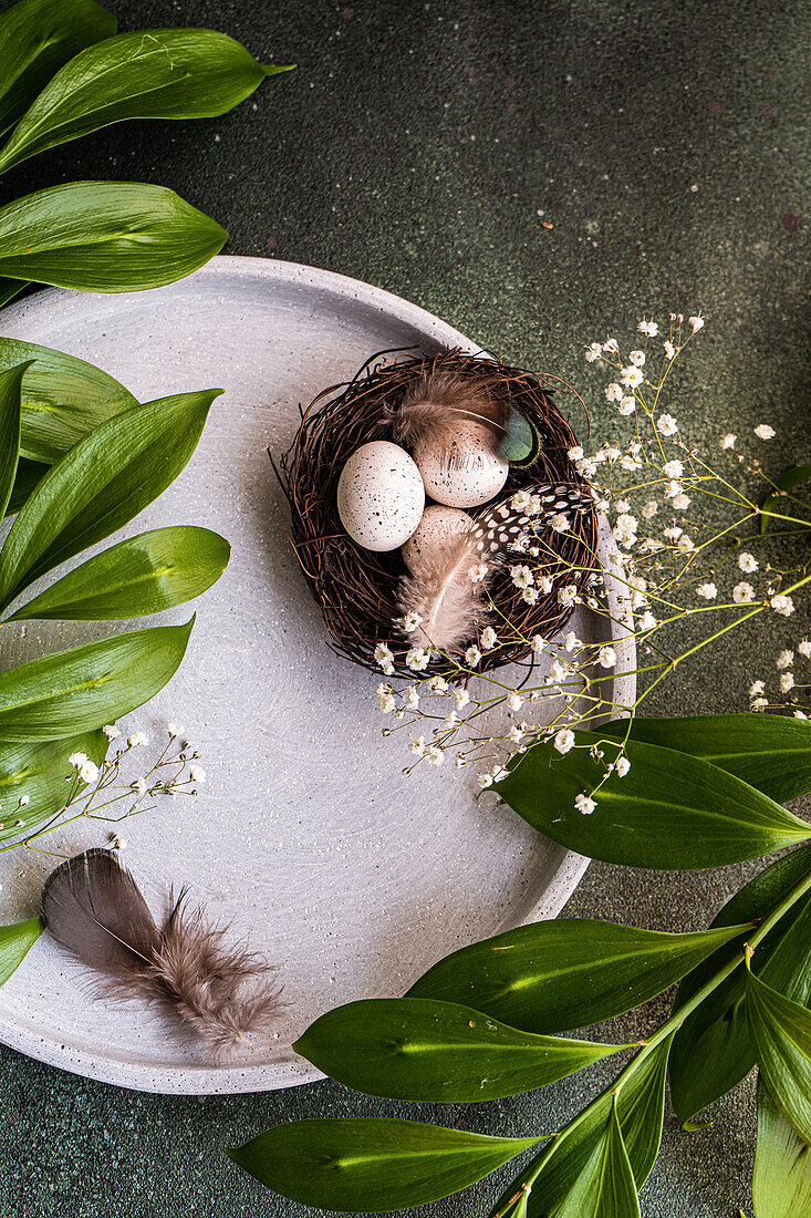 From above place setting for Easter dinner with ceramic plates easter eggs in nest surrounded by Italian ruscus leaves