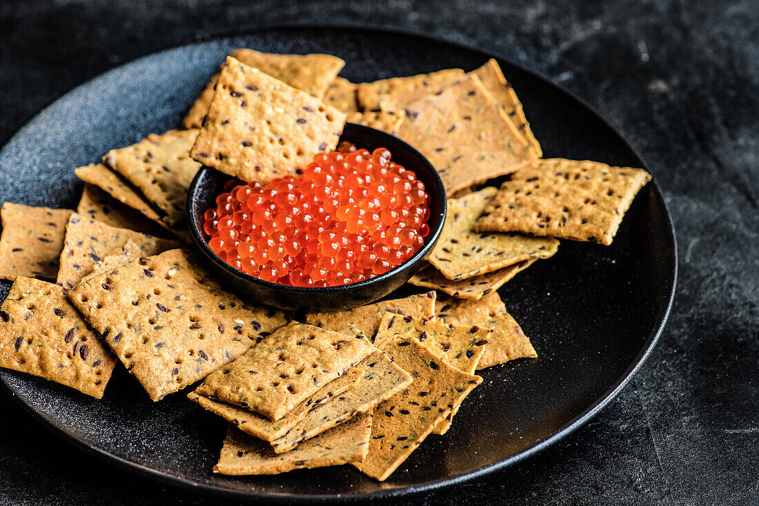 Red fresh trout fish caviar in a bowl served with sesame seed crackers on concrete table background