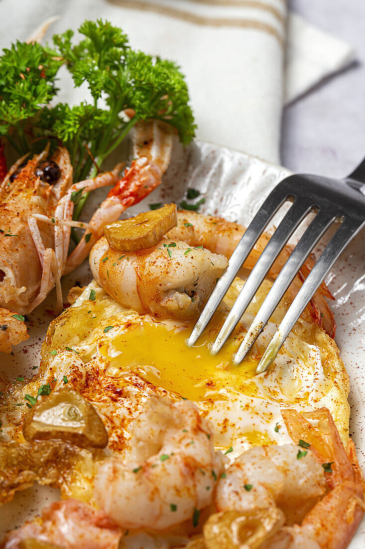 From above appetizing fried eggs with king prawns served on white plate with green herbs and seasoning on towel in light kitchen