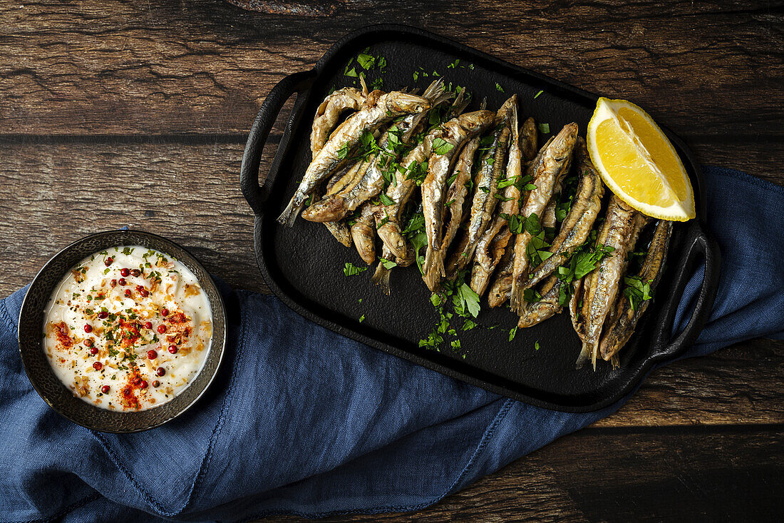 Top view of tasty fried anchovies with juicy lemon piece and chopped parsley on tray against savory sauce