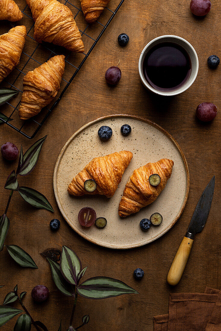 From above of tasty fresh baked croissants served on plate with fruits placed near cup of tea on wooden table in morning time in light room