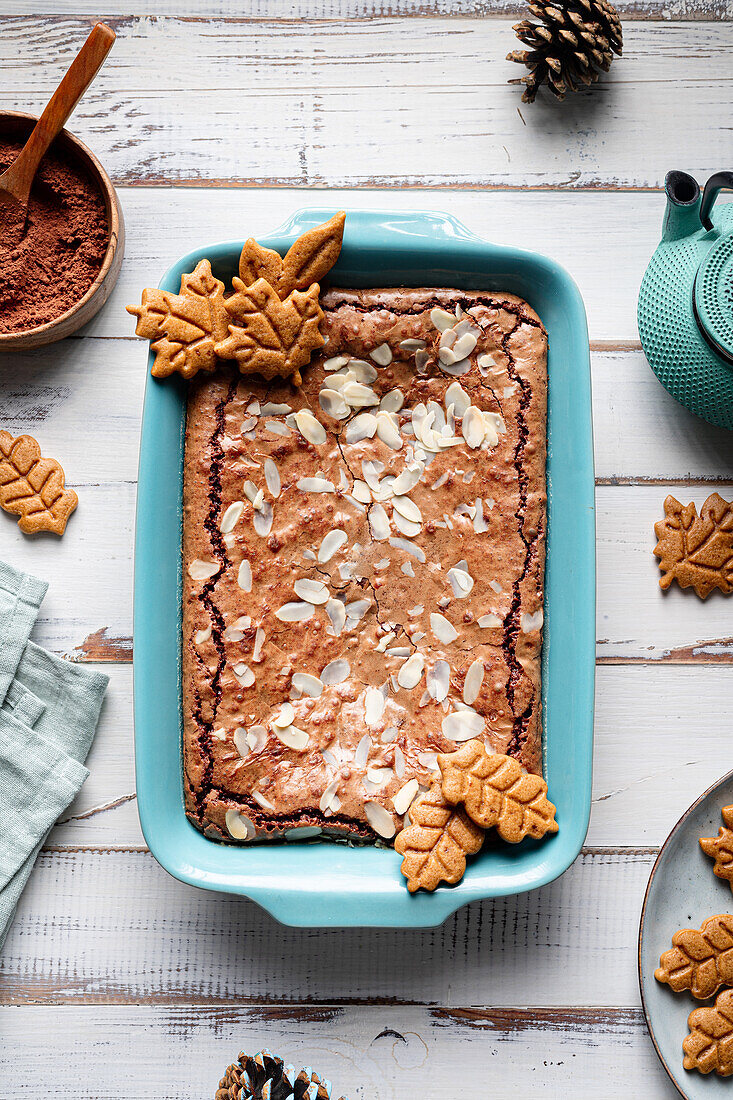Draufsicht auf einen leckeren gebackenen Brownie mit Mandelblättchen in einem Backblech mit Keksen auf einem Holztisch mit Kakaopulver