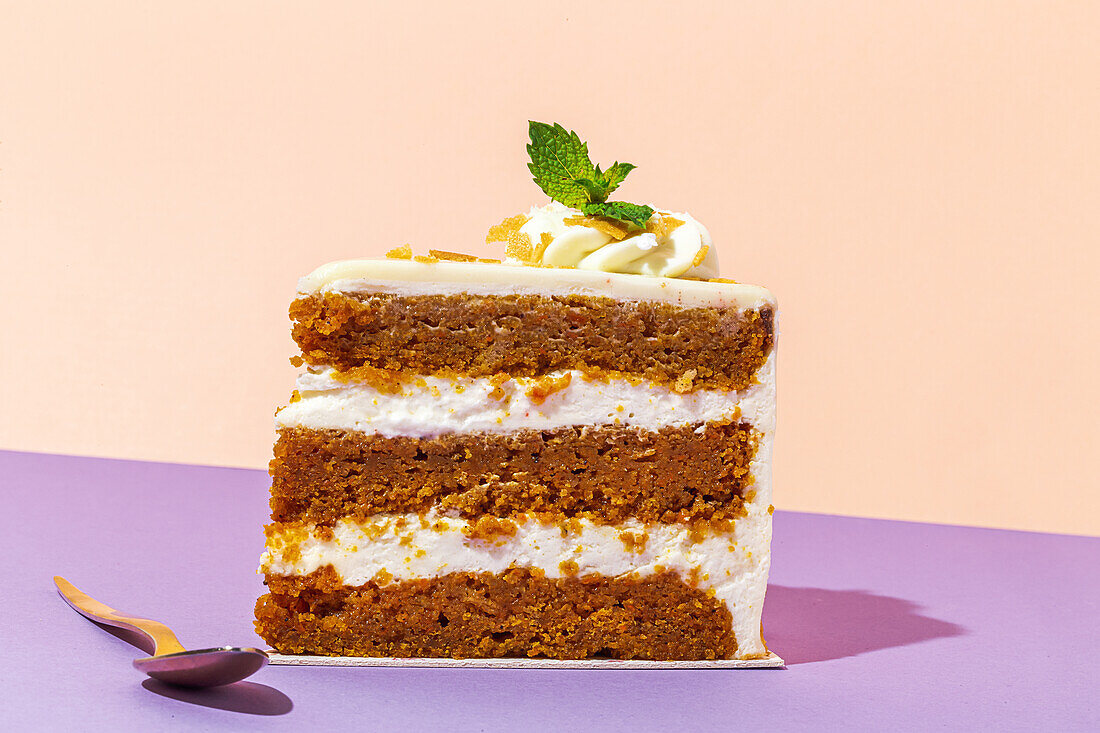 Slice of tasty sweet carrot sponge cake with cream decorated with mint leaf served on plate with spoon on table on colorful background