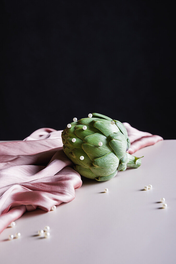 Composition of fresh healthy artichoke with small beads placed on draped fabric on white table and black ground