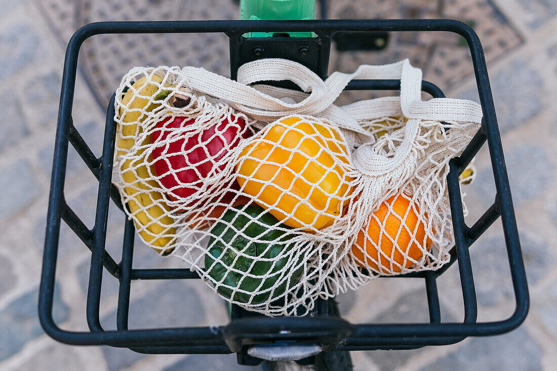 Umweltfreundlicher Netzbeutel mit frischem Obst und Gemüse im Metallkorb eines geparkten Fahrrads in der Stadt (von oben)