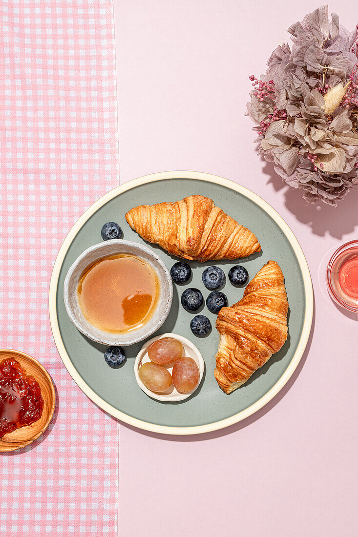 Top view of delicious croissants served on ceramic plate with fresh blueberries and jam placed on pink table