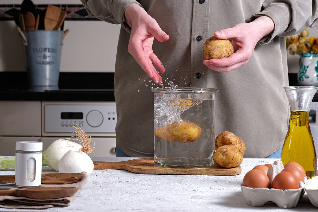 Anonyme Person schüttet rohe, ungeschälte Kartoffeln in ein Glas mit Wasser, während sie in der Küche Zutaten für ein hausgemachtes spanisches Omelett vorbereitet