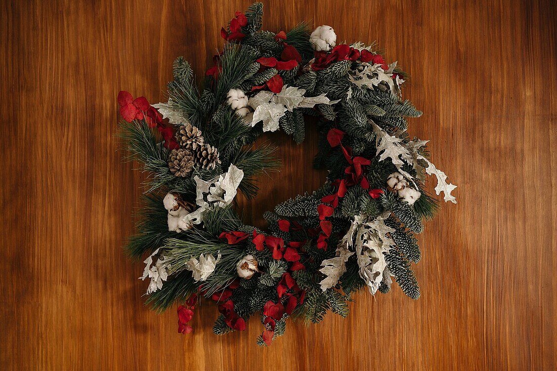 Stylish Christmas wreath with coniferous twigs and decorative elements hanging on wooden wall in daylight