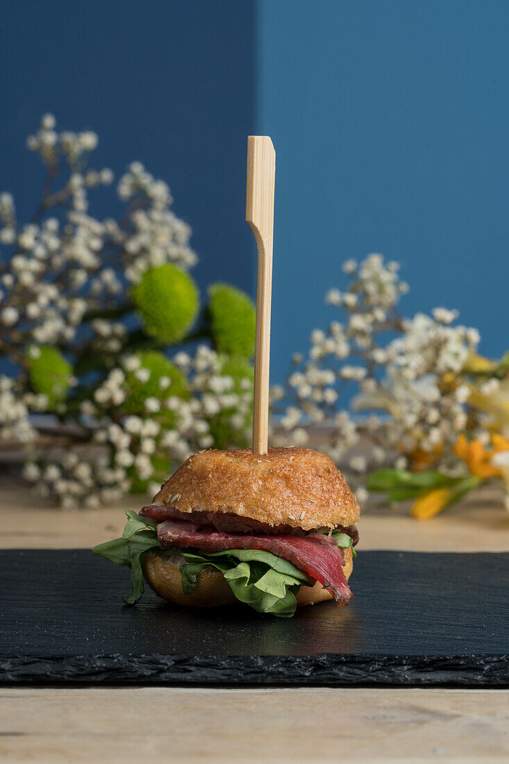 Frischer appetitlicher Mini-Burger auf schwarzem Brett neben blühenden Blumen vor blauer Wand serviert