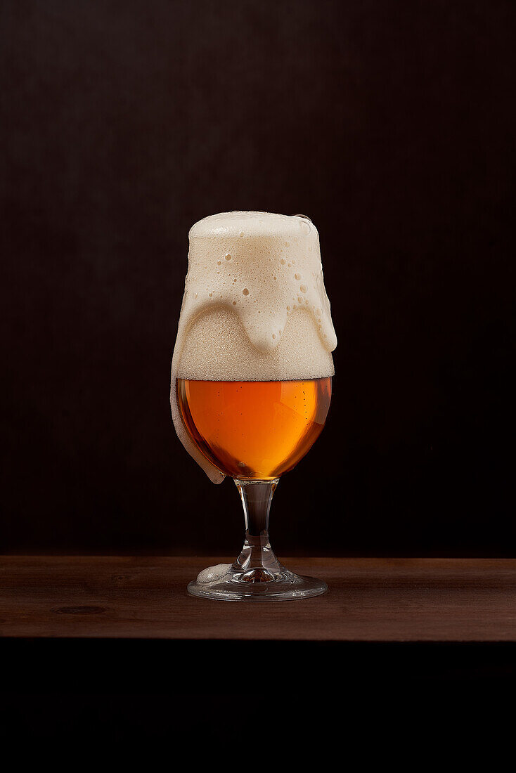 Freshly brewed foamy beer in tulip glass placed on wooden counter against brown background