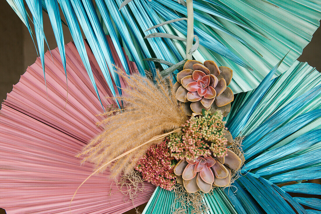 Kreative Komposition aus frischen Sukkulenten mit zarten Blüten und trockenem Schilf, arrangiert mit bunten Palmenblättern einer exotischen Pflanze bei Tag