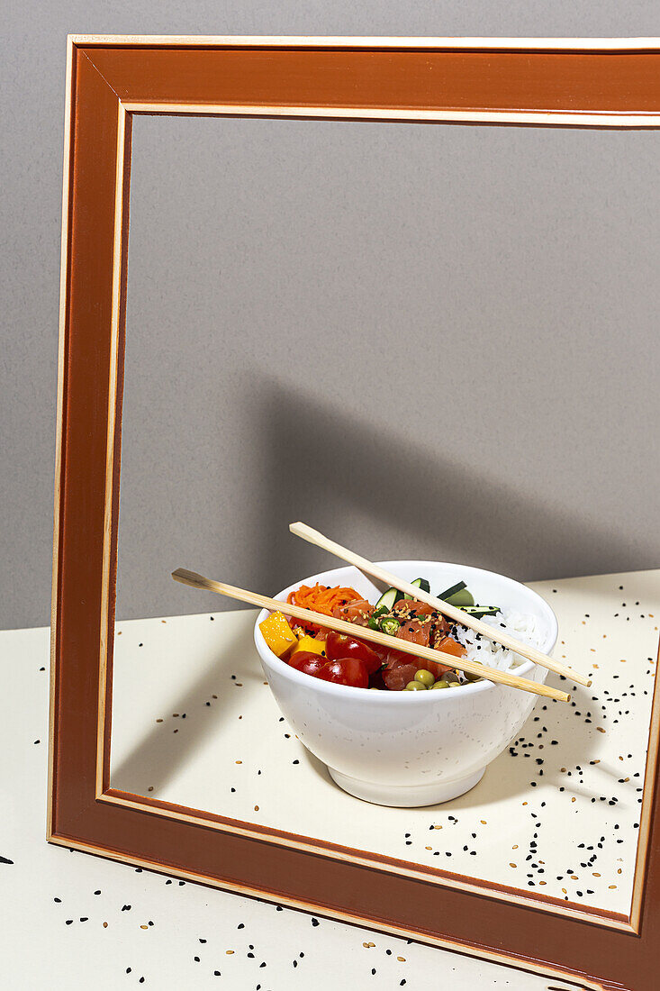 High angle of white bowl with tasty poke dish and chopsticks placed behind frame on table covered with sesame seeds