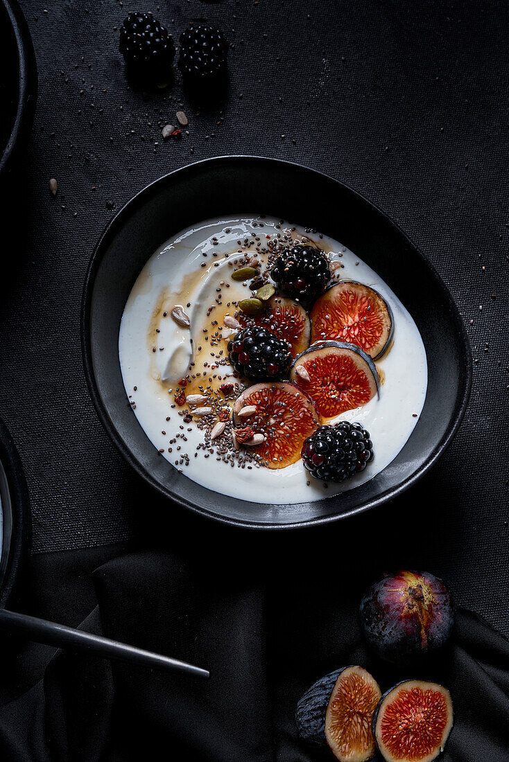 Appetitanregendes gesundes Frühstück bestehend aus Joghurt, frischen Feigen und Brombeeren, bestreut mit Samen und Nüssen auf einem schwarzen Tisch von oben