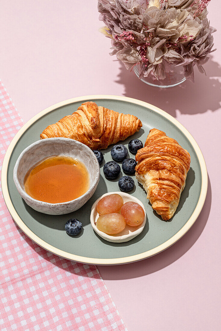 Leckere Croissants auf einem Keramikteller mit frischen Blaubeeren und Marmelade auf einem rosa Tisch