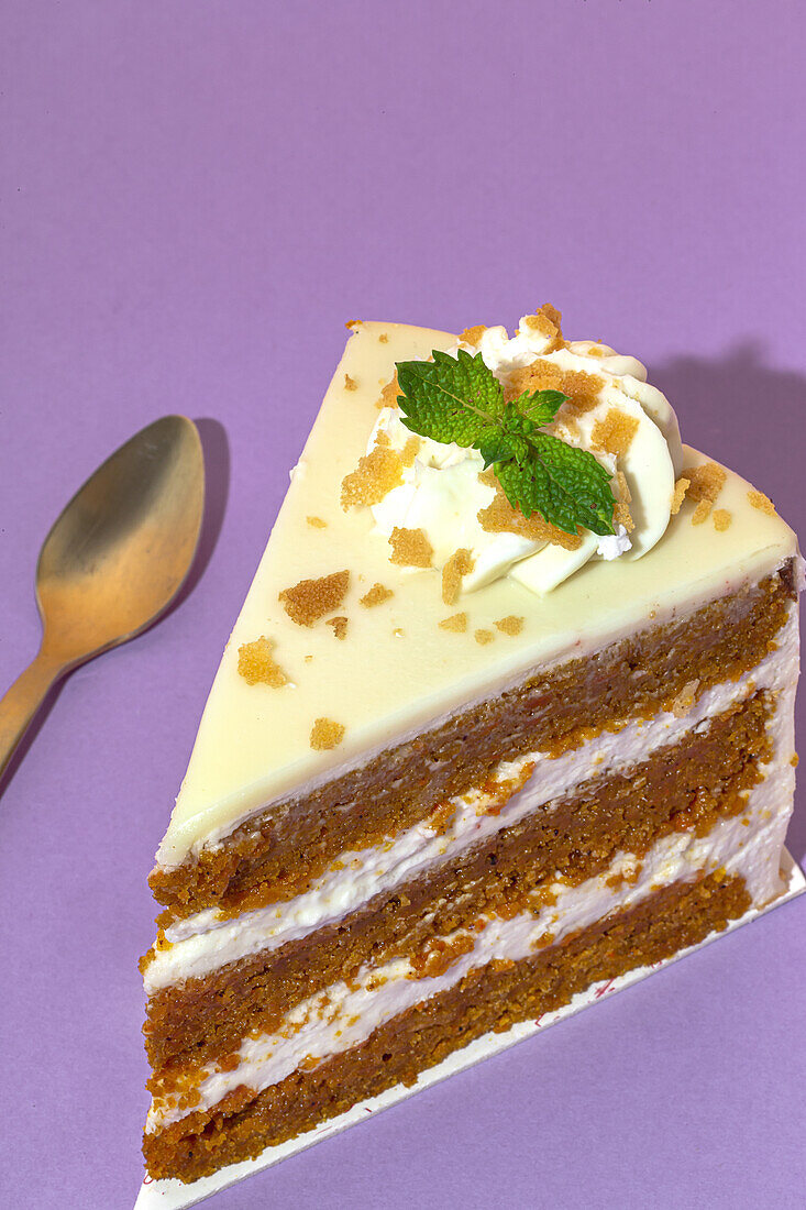 From above close up of slice of tasty sweet carrot sponge cake with cream decorated with mint leaf served on plate with spoon on table on purple background