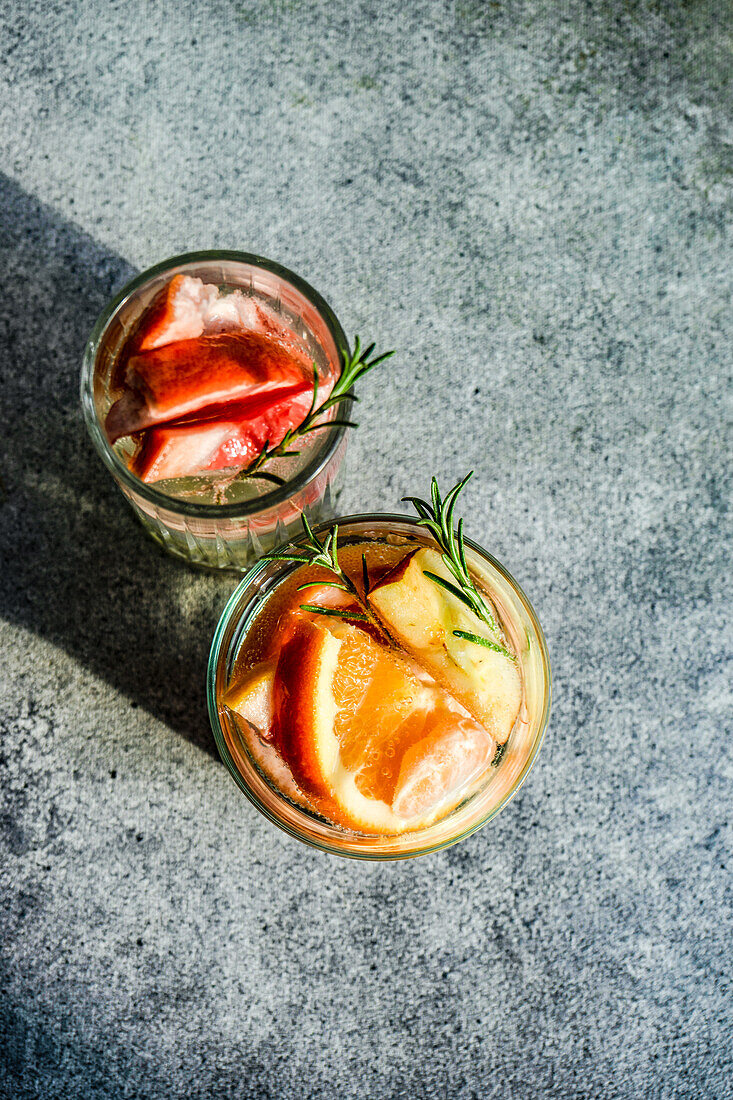 Draufsicht auf einen tropischen Sommercocktail aus Wodka und Sprite mit reifen Früchten und Rosmarin auf grauem Hintergrund