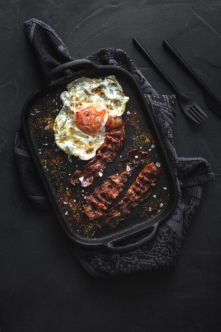 Top view of sunny side up egg with fried bacon slices and condiments on tray against cutlery on dark background