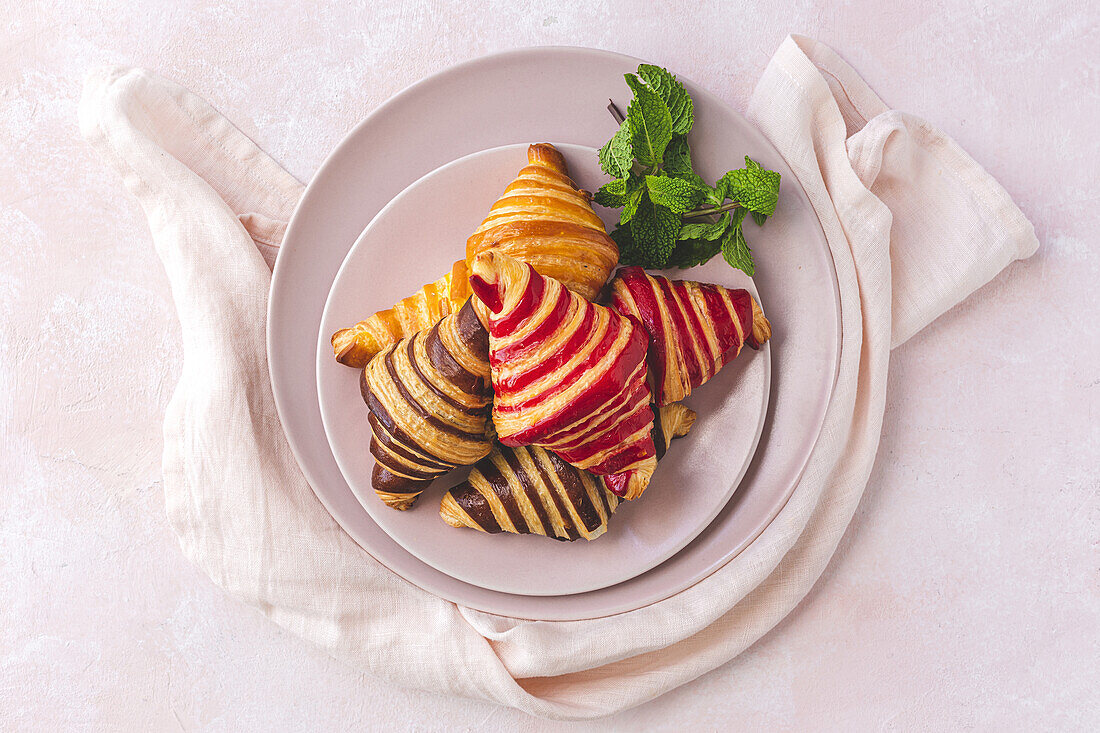 Draufsicht auf leckere Croissants auf einem Teller mit einem Minzzweig auf pastellrosa Hintergrund