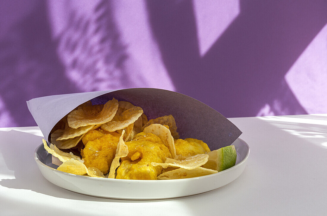 Appetizing fried chips and fish in paper wrapping served on plate on table with bright sunlight in cafe