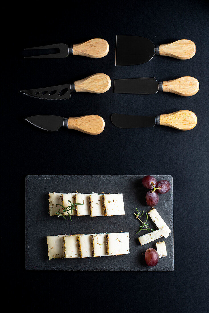 Draufsicht auf verschiedene Messer in der Nähe von leckerem Käse und Trauben auf schwarzem Brett