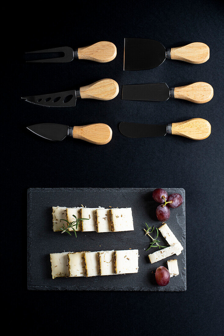 Top view of various knives placed near delicious sliced cheese and grape on black board