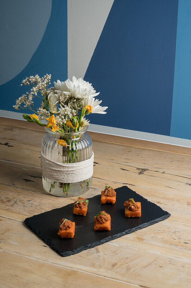 From above of delicious appetizer in square bites served on black board near bouquet of blooming flowers in glass vase against blue wall