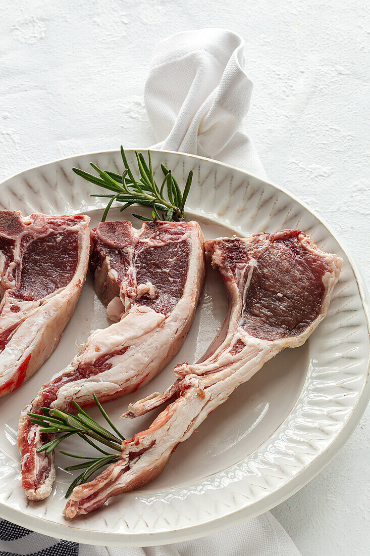 From above of uncooked lamb chops served on white ceramic plate with fresh green rosemary on table in light kitchen