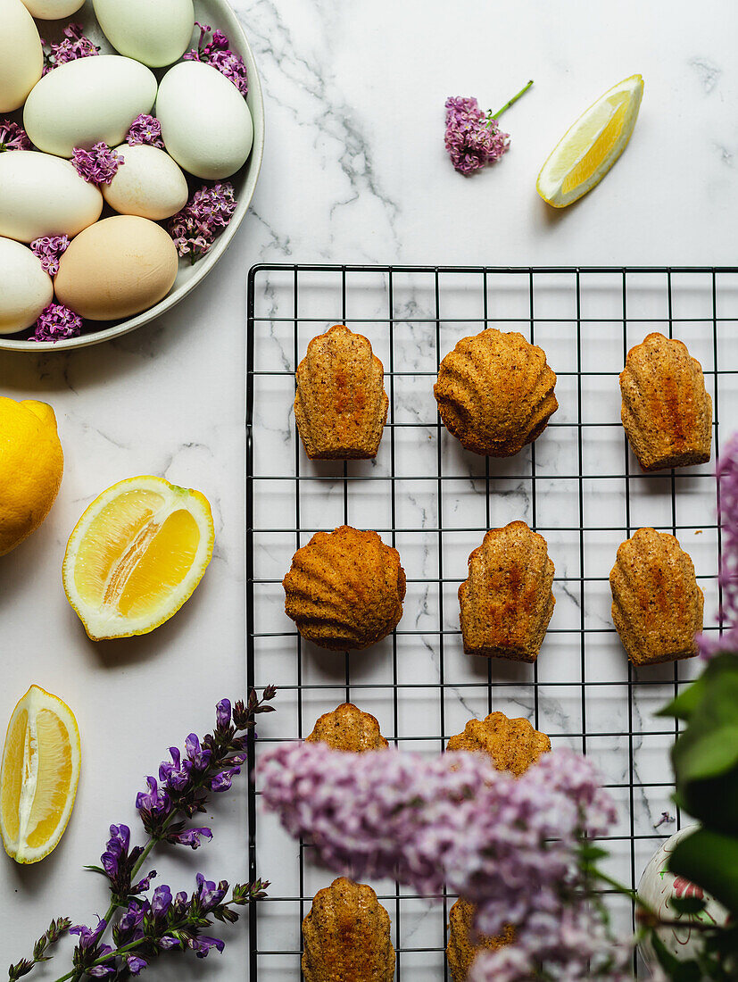 Top view of tasty madeleines on cooling rack near plate with eggs and lavender flowers on marble surface