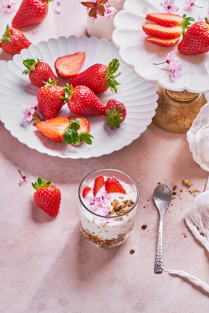 From above of glass of delicious yogurt with granola placed on table near plates of fresh ripe strawberries served on table in kitchen