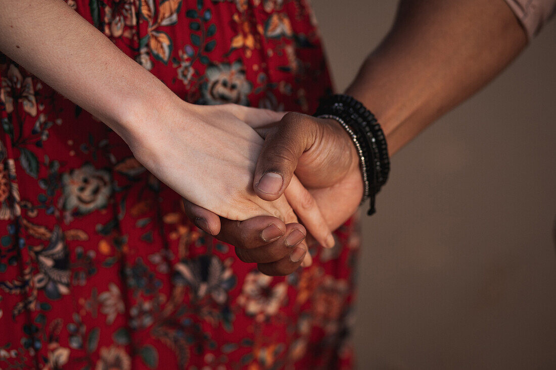 Anonyme Frau in buntem Kleid und schwarzer Mann mit Armband am Handgelenk halten sich an den Händen und genießen romantische Momente miteinander