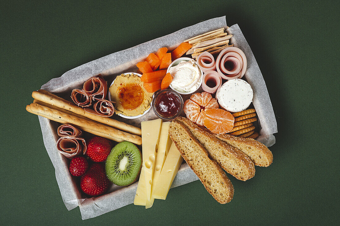 Von oben Brunch-Box mit verschiedenen Wurst- und Käsesorten und Knäckebrot in der Nähe von reifen Kiwis, süßen Erdbeeren und geschälten Mandarinen in der Nähe von Marmelade in einem Glas auf einem Holztisch in der Nähe einer Serviette