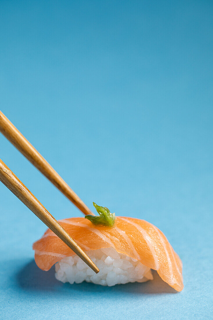 Leckeres japanisches Nigiri mit frischem Lachs und grünem Wasabi vor blauem Hintergrund mit Stäbchen in einem hellen Studio