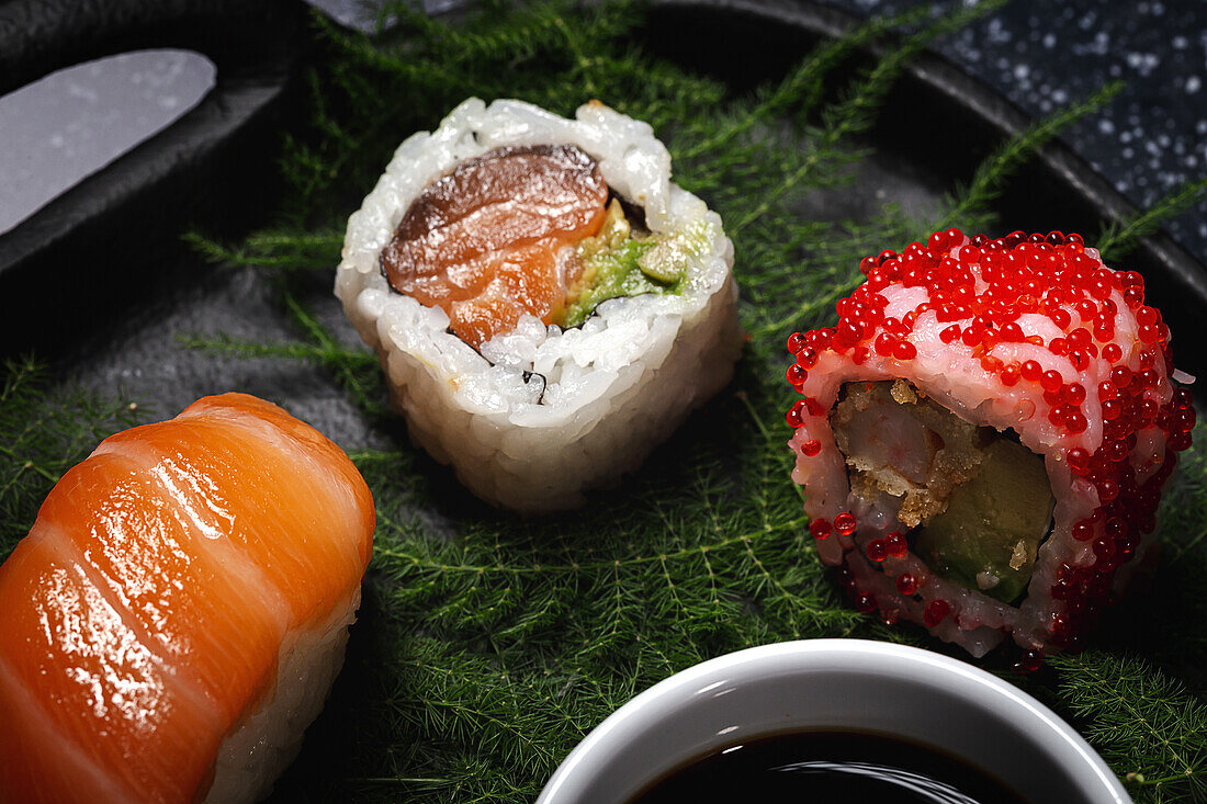 Tasty fresh assorted sushi served on green plant twigs on black plate with soy sauce on marble table near chopsticks