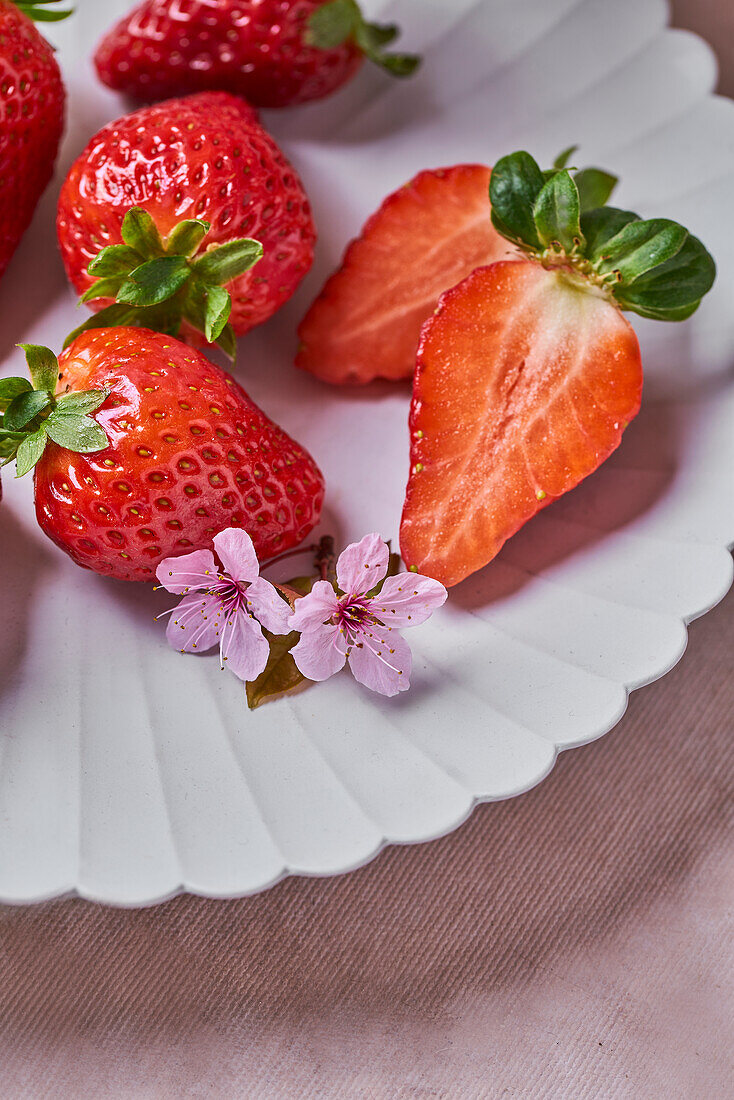 Frische reife ganze und geschnittene Erdbeeren und zarte rosa Blume auf weißem Teller auf dem Tisch
