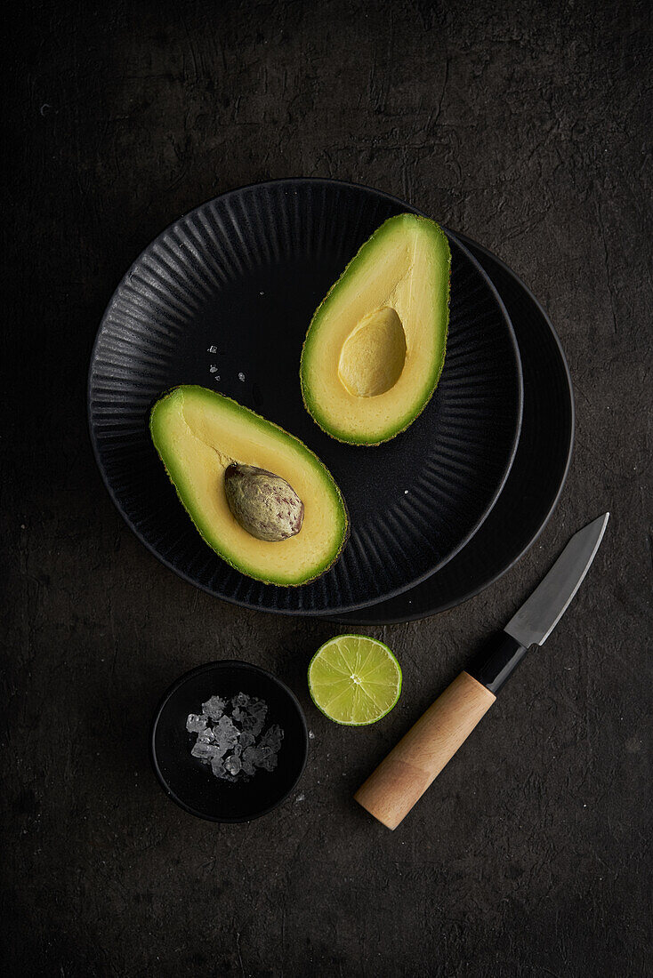 Frische Avocadohälften von oben auf einem Tisch mit grüner Limette, Salz und Messer auf dunklem Hintergrund angeordnet